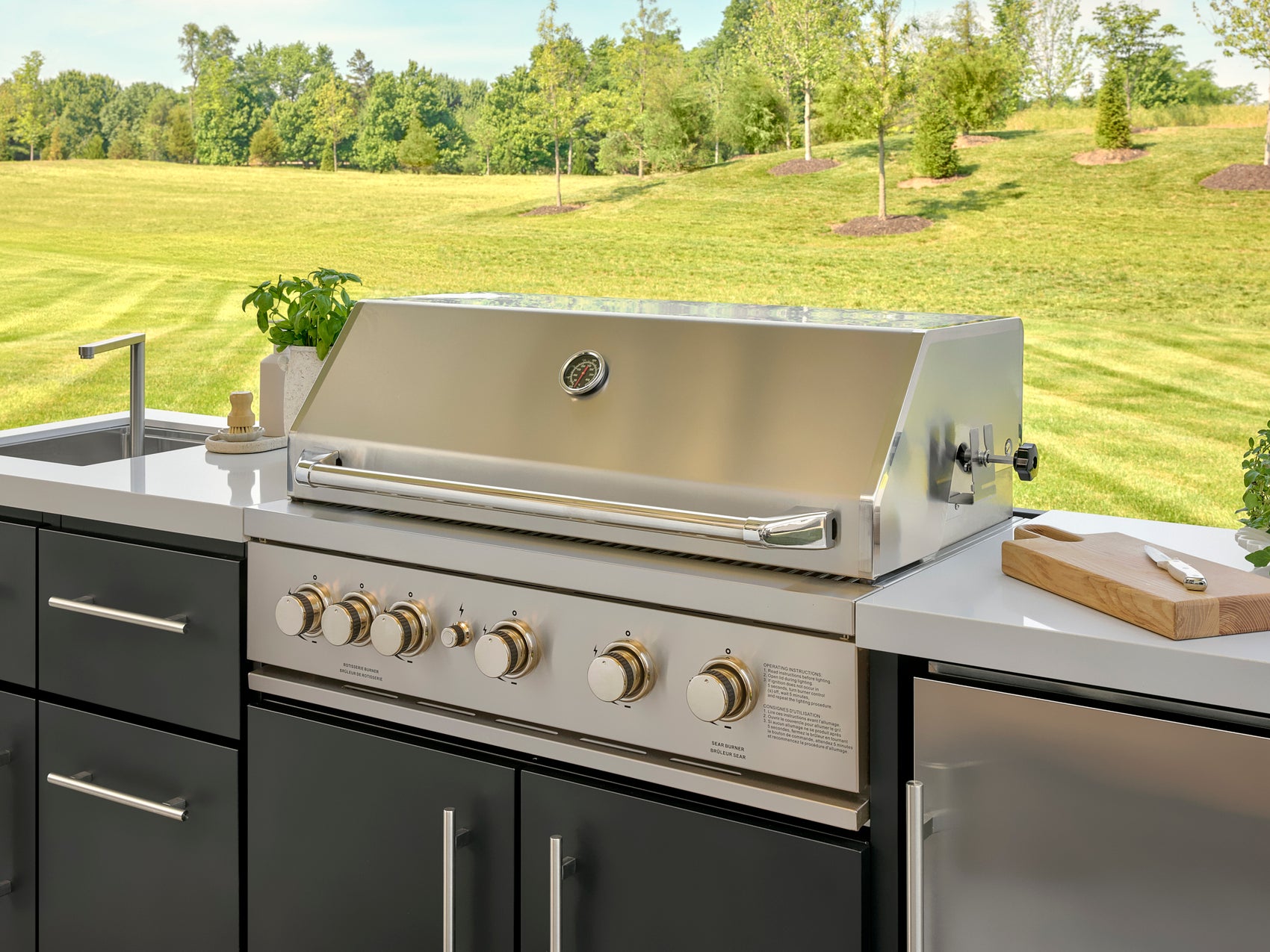 Stainless steel Emory outdoor grill with temperature gauge and control knobs built into cabinetry with white countertop.