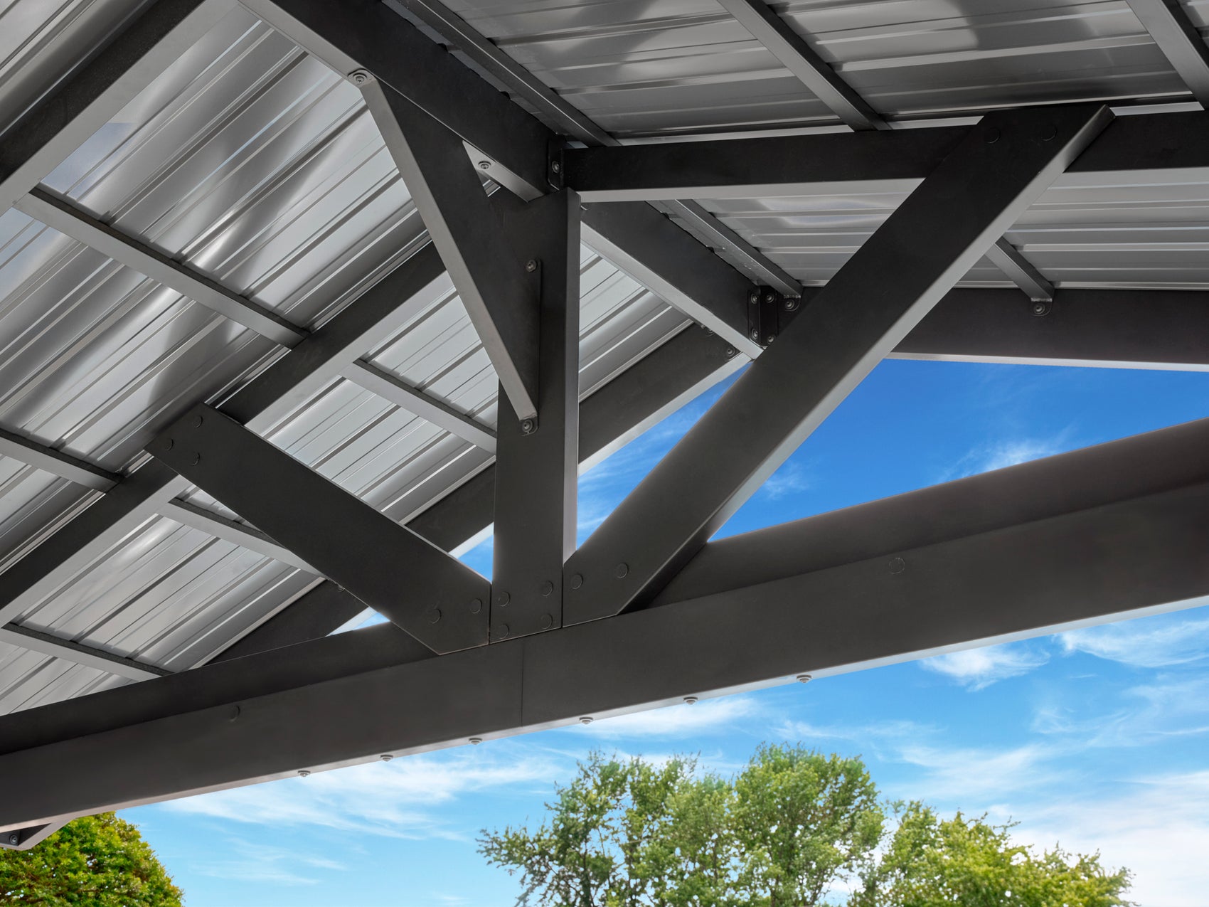 Steel Lancaster outdoor gazebo with beams, corrugated metal roof panels, and diagonal bracing under blue sky with trees.