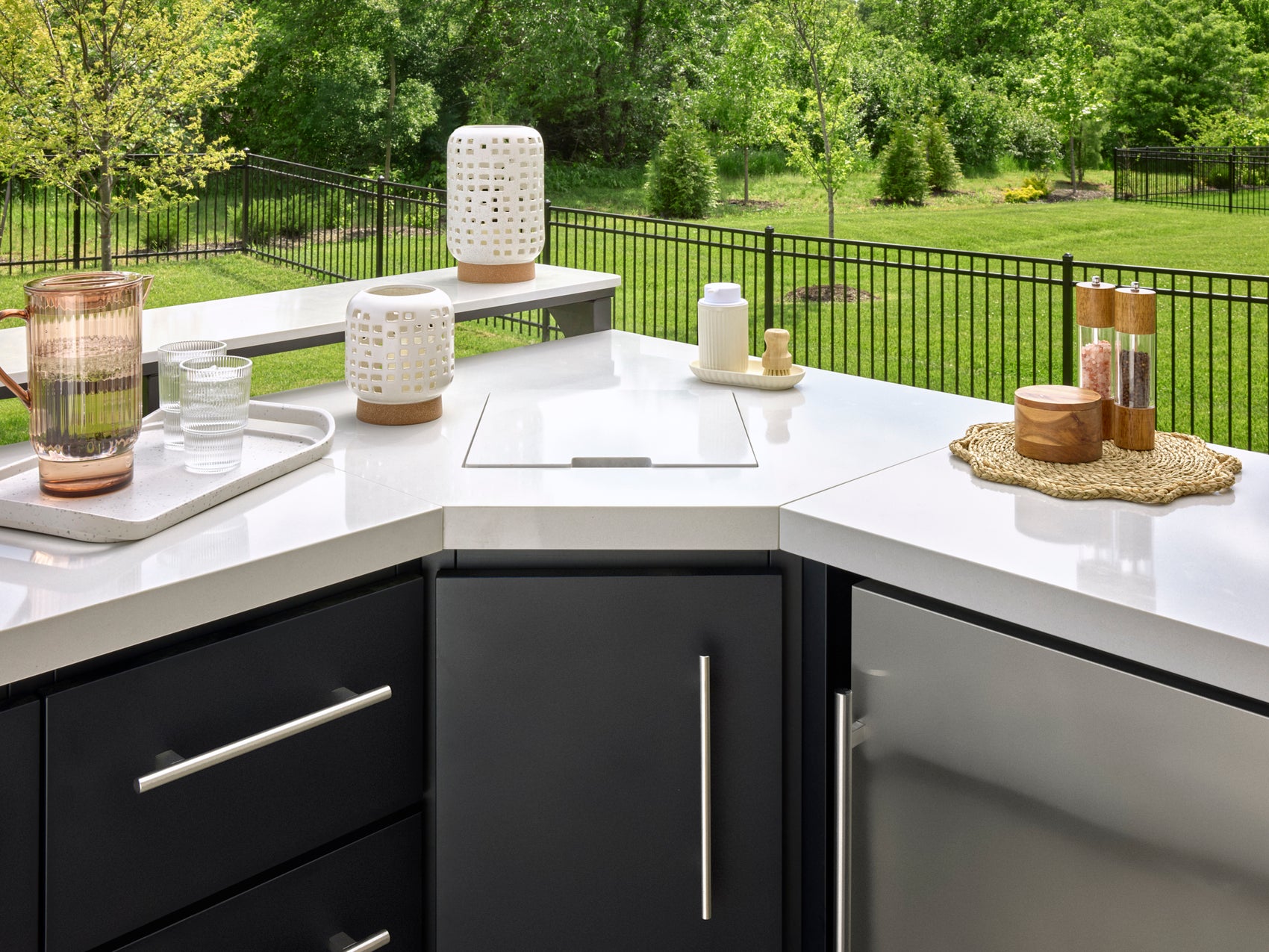Built-in sink with cover closed on white engineered stone countertop with black cabinets overlooking fenced backyard.