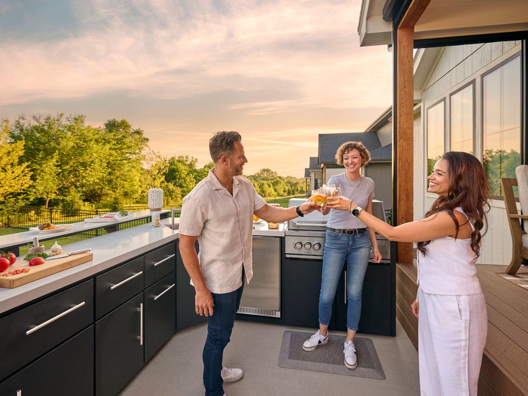 Three people toasting with drinks on deck with outdoor kitchen with dark cabinetry and built-in grill under patio pergola.