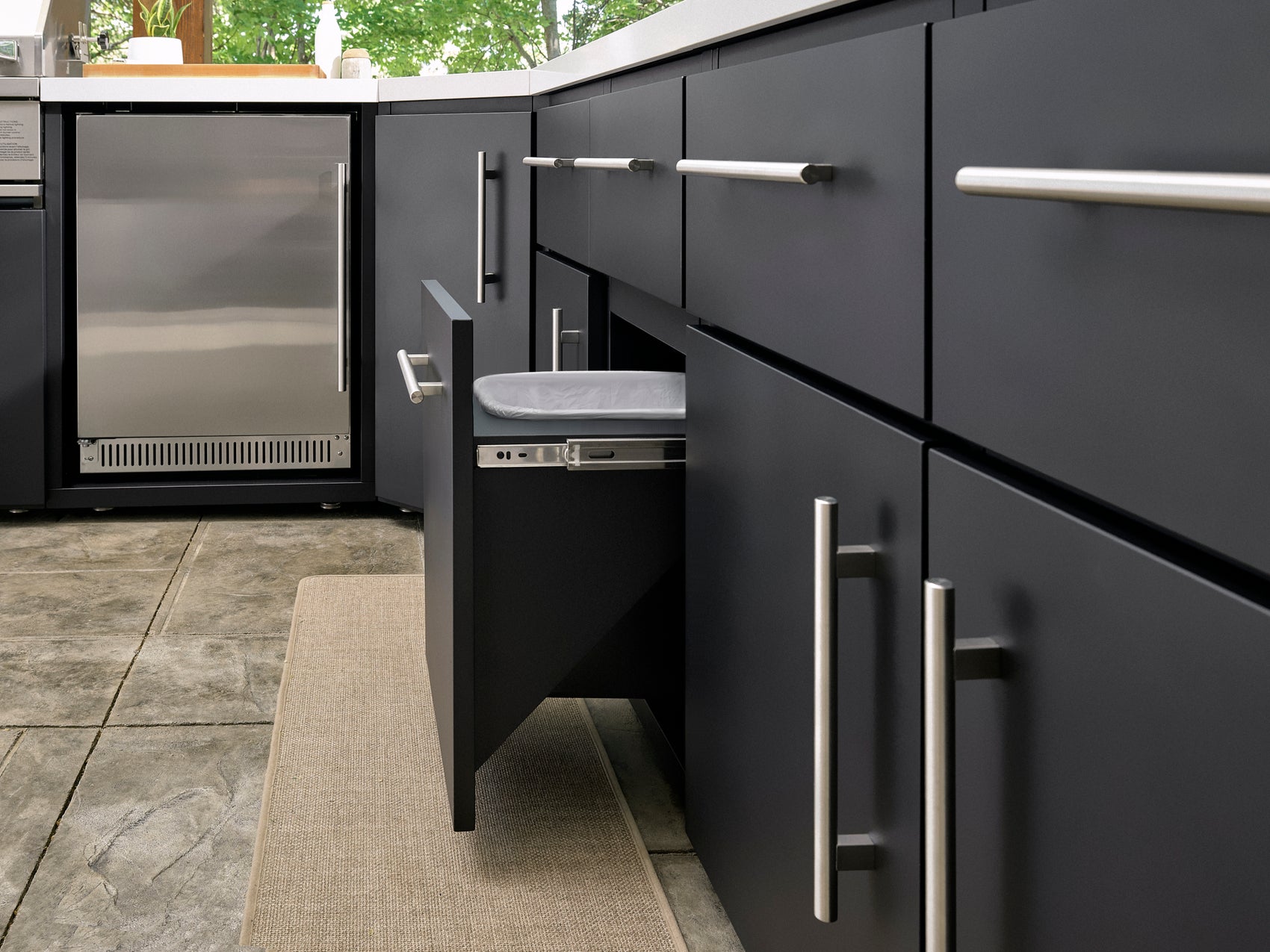 Emory modern outdoor kitchen with matte black cabinetry, pull-out trash drawer, built-in refrigerator, and stone flooring.