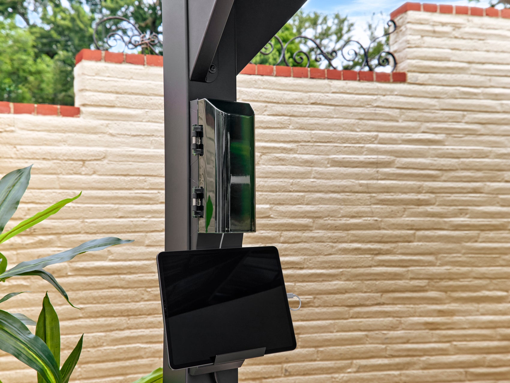 Steel patio gazebo column with integrated tablet mount and electrical outlets against textured stone wall and greenery.