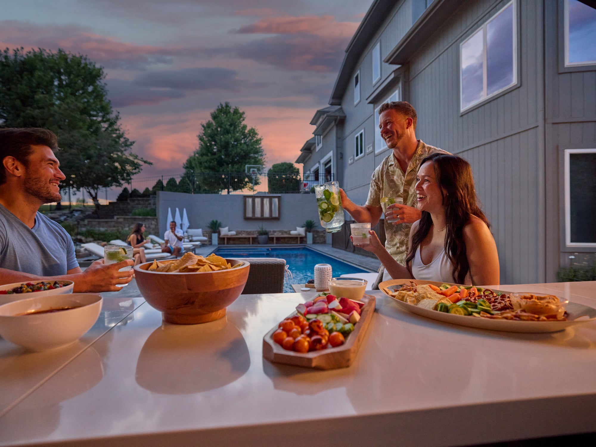 Friends enjoying outdoor dining at Range Outdoor Living Lancaster kitchen in backyard by a pool at sunset.