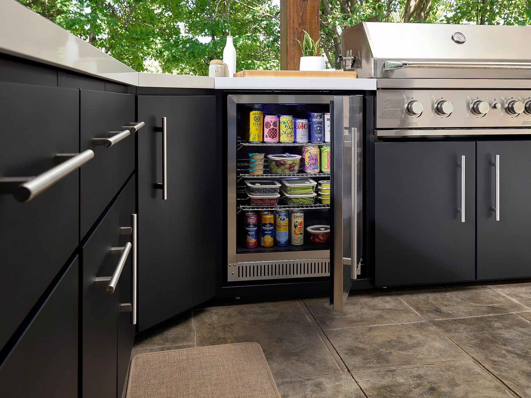 Open glass-door built-in refrigerator stocked with beverages and food containers in Emory outdoor kitchen with cabinetry.