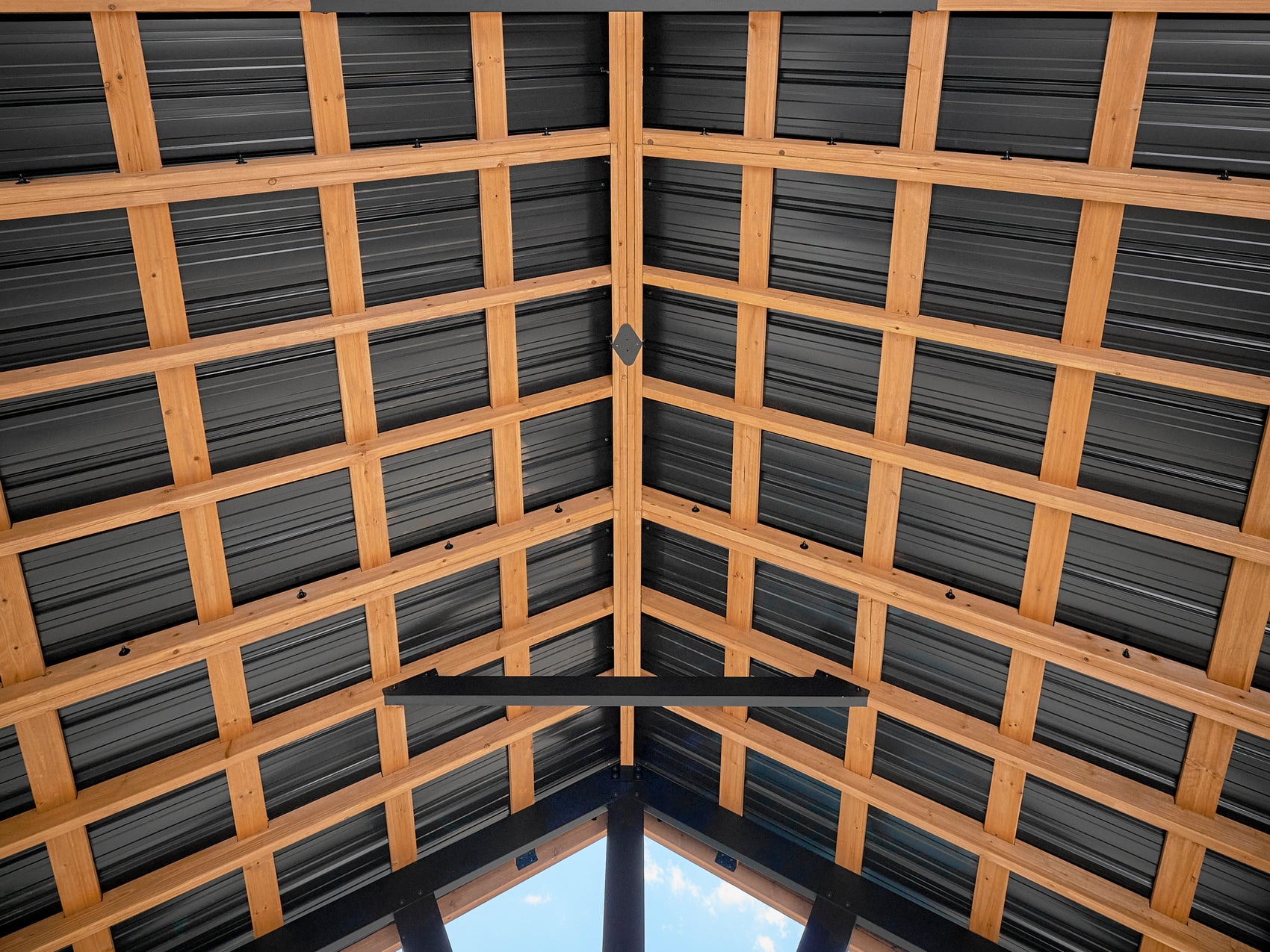 Interior ceiling view of Lodge gazebo wooden beam framework with dark corrugated metal roofing panels and center ridge.