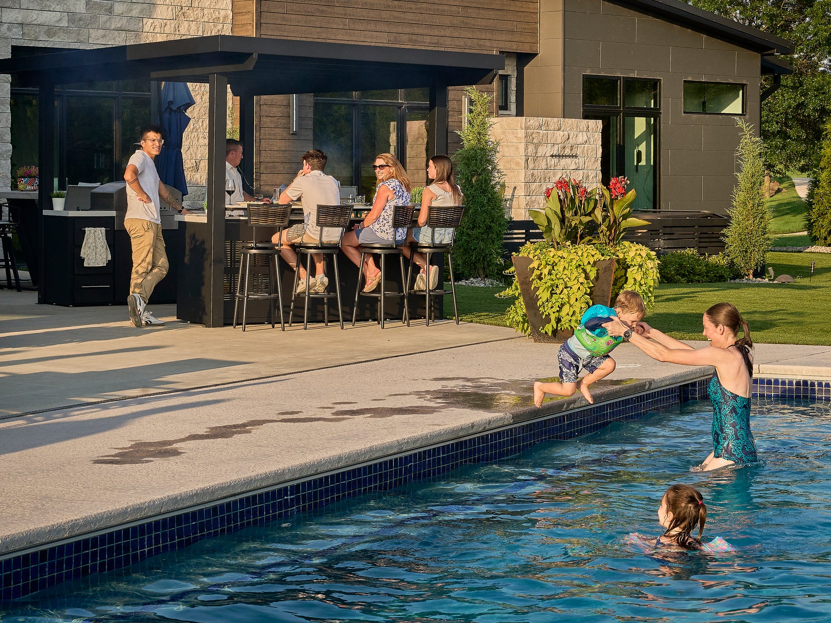 Family playing in pool while others sit at bar seating under galvanized steel roof of Edgemont Bay outdoor kitchen.