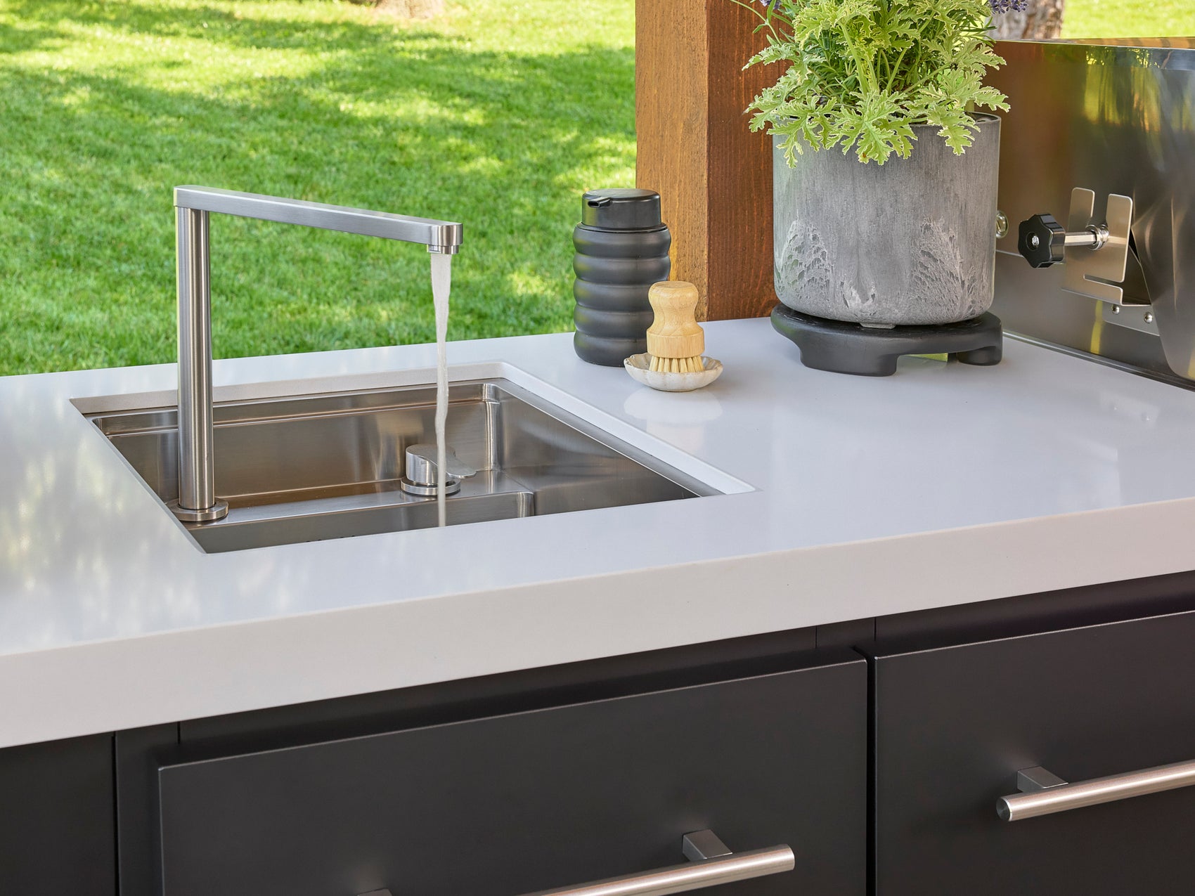 Modern stainless steel sink with tall faucet set in white engineered stone countertop with black cabinets and greenery.