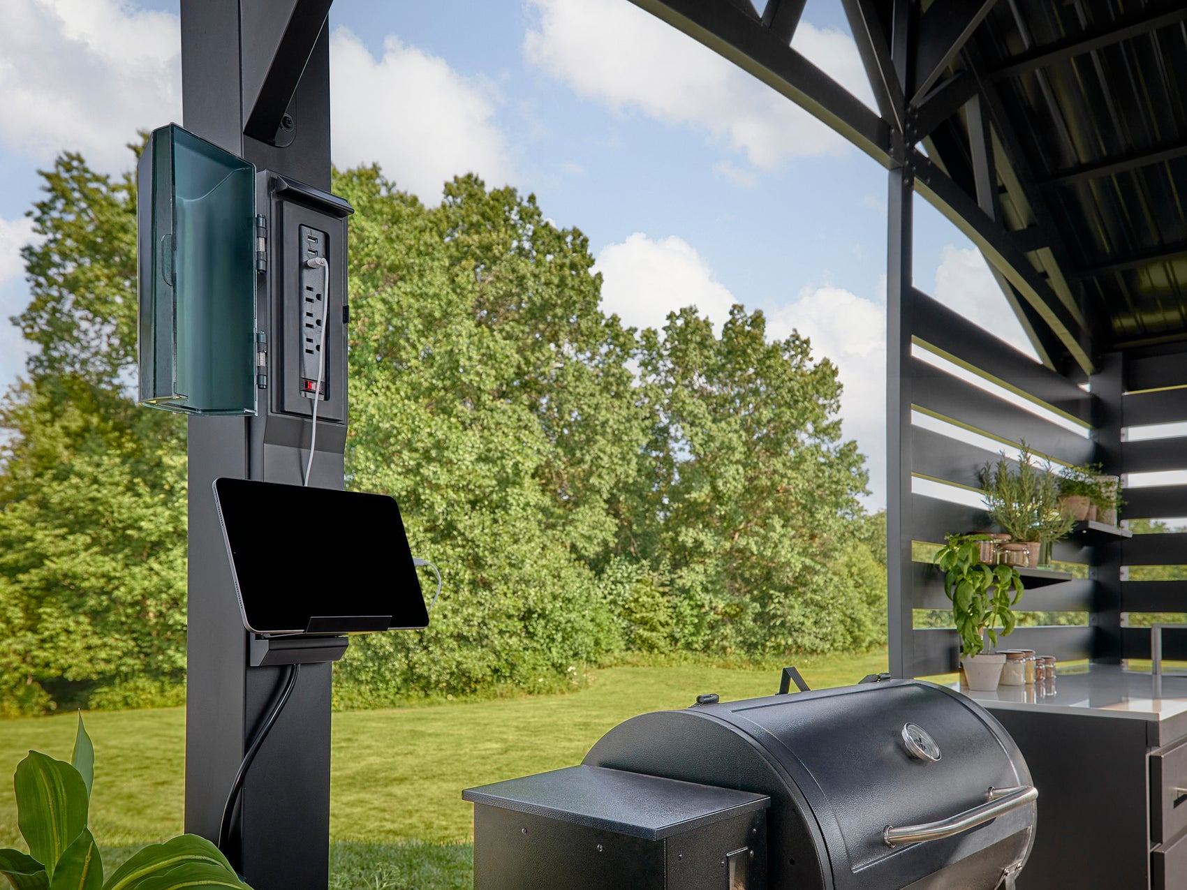 Outdoor electrical outlet and panel mounted on backyard kitchen bracket beside stainless steel 5-burner grill.