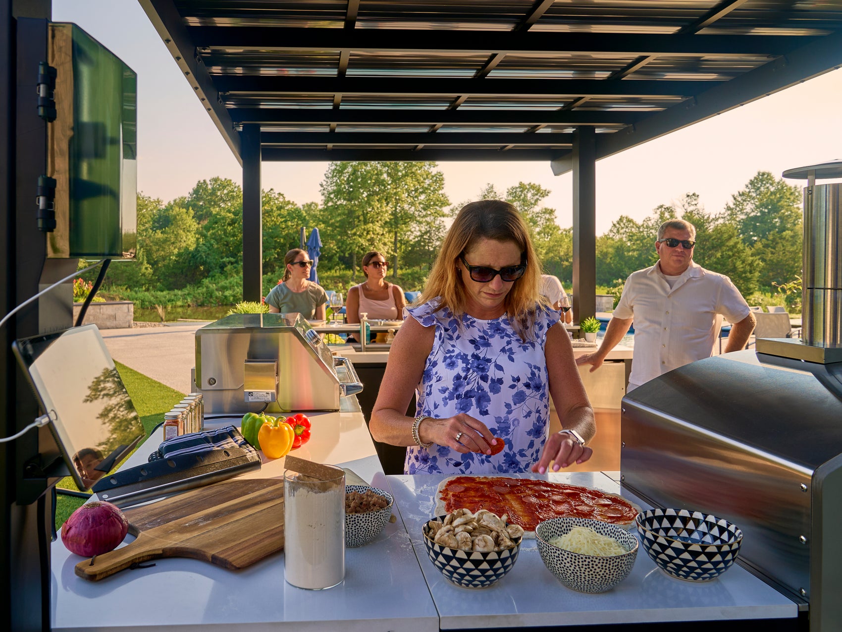 Family gathering at Edgemont Bay outdoor kitchen island with woman prepping pizza on engineered stone counter.