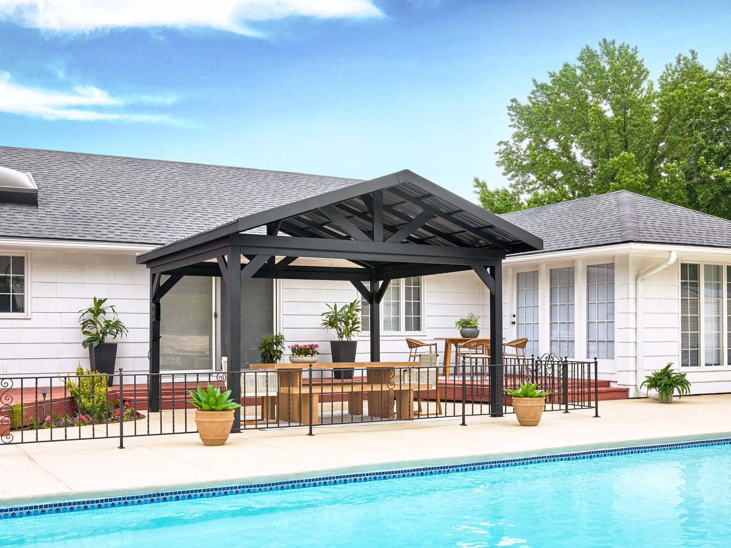 Dark gray Lancaster hardtop gazebo with dining table beside pool and white house with landscaping under blue sky.
