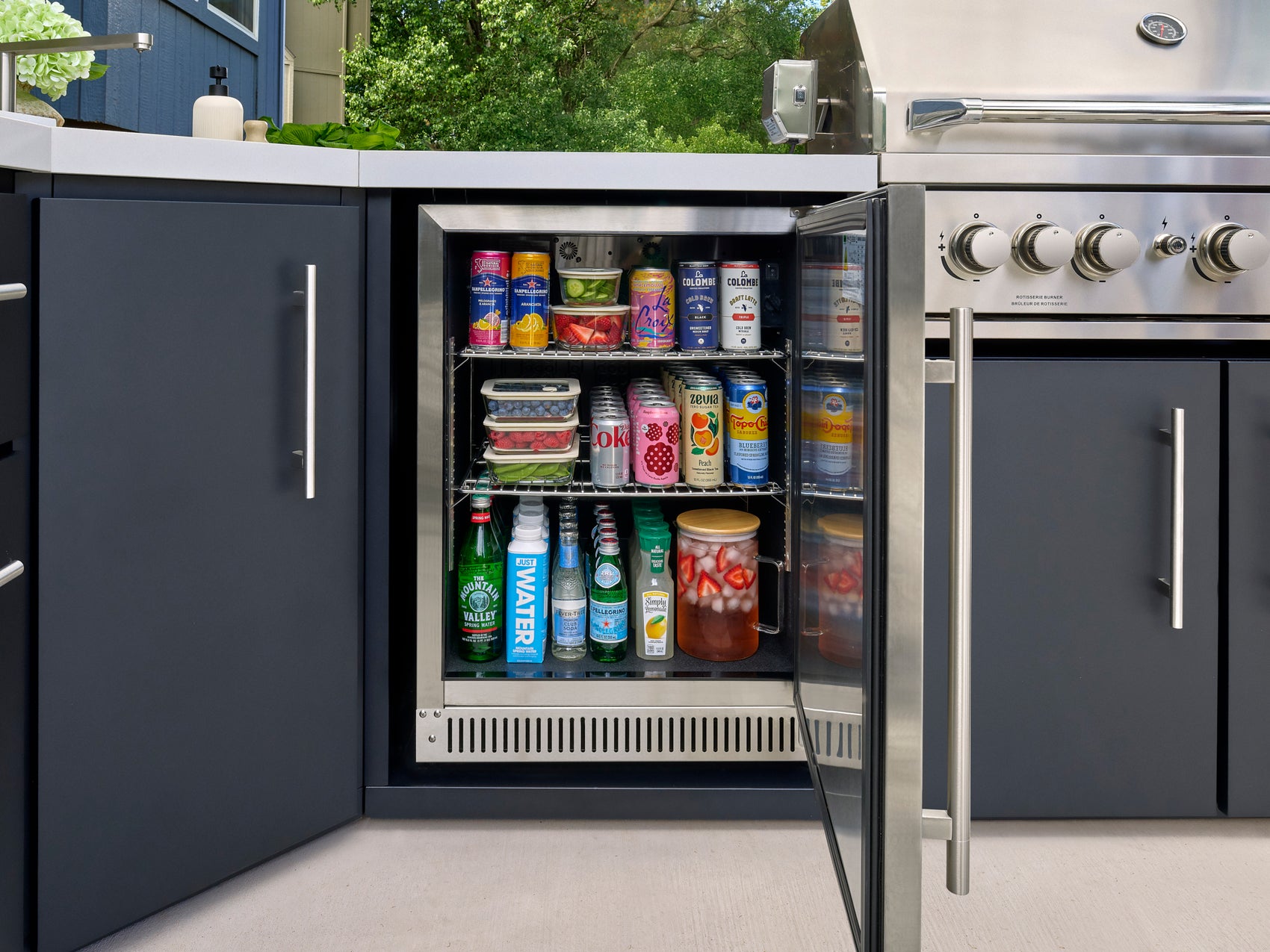 Outdoor beverage cooler with cans, bottles, and food containers on three wire shelves in dark gray Emory outdoor kitchen.