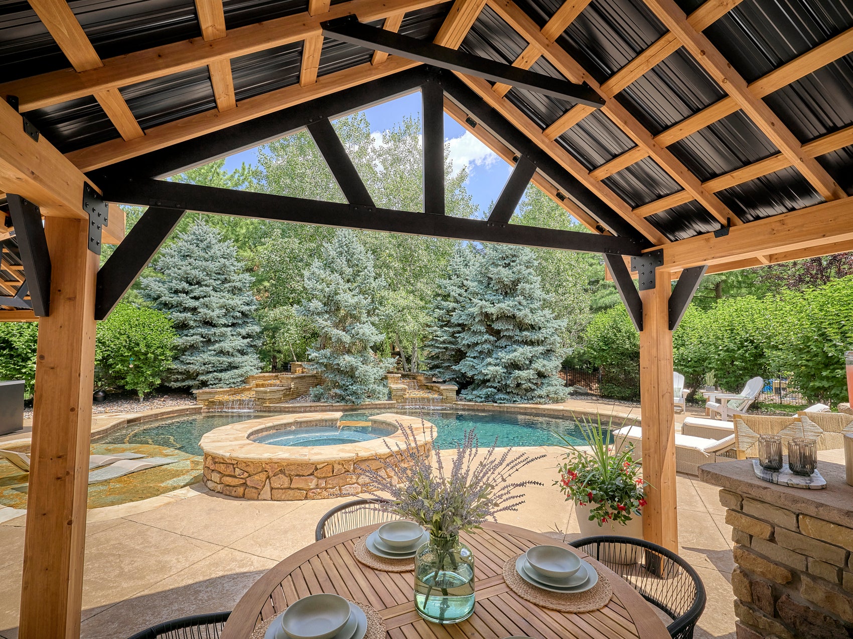 Lodge garden gazebo with metal roof and wood beams overlooking pool, spa, stone landscaping, and outdoor seating area.