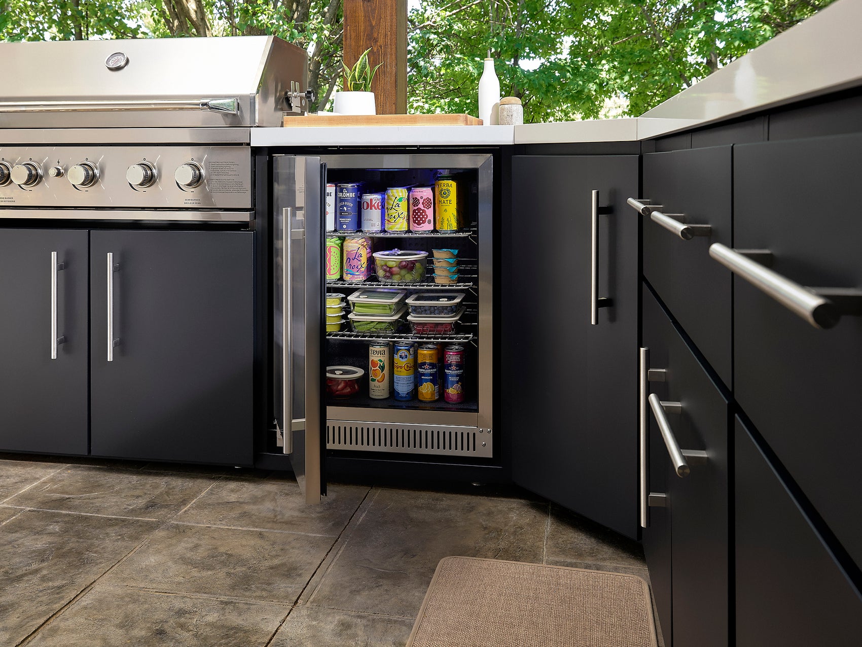 Open outdoor mini fridge with stocked with beverages and food containers in dark cabinetry beside stainless grill.