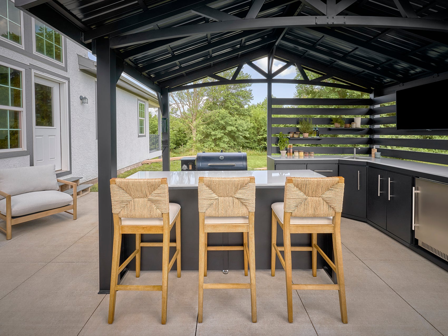 Outdoor patio bar with engineered stone top, woven counter stools, black cabinets, and grill under covered pergola.