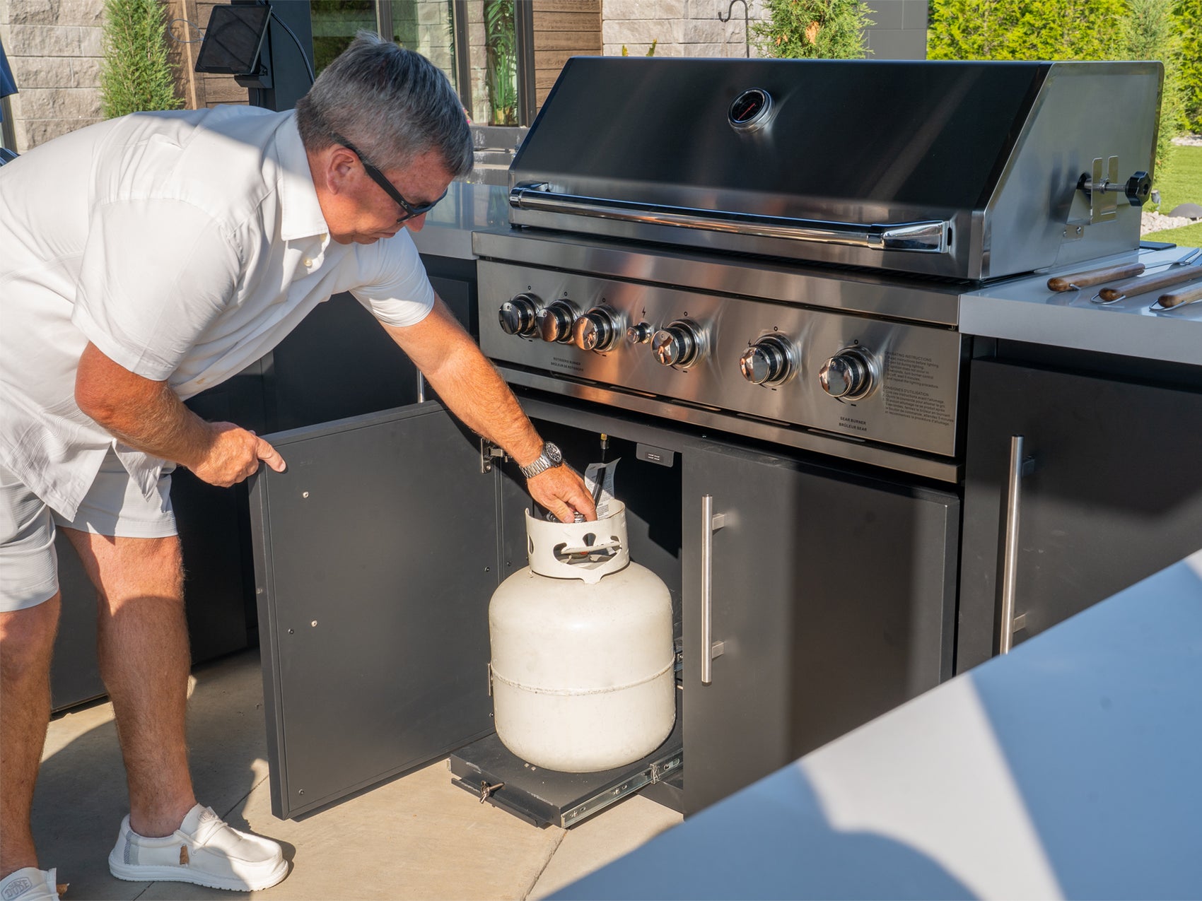 Man accessing slide-out propane drawer under Range Outdoor Living 5-burner grill on Edgemont Bay backyard kitchen.