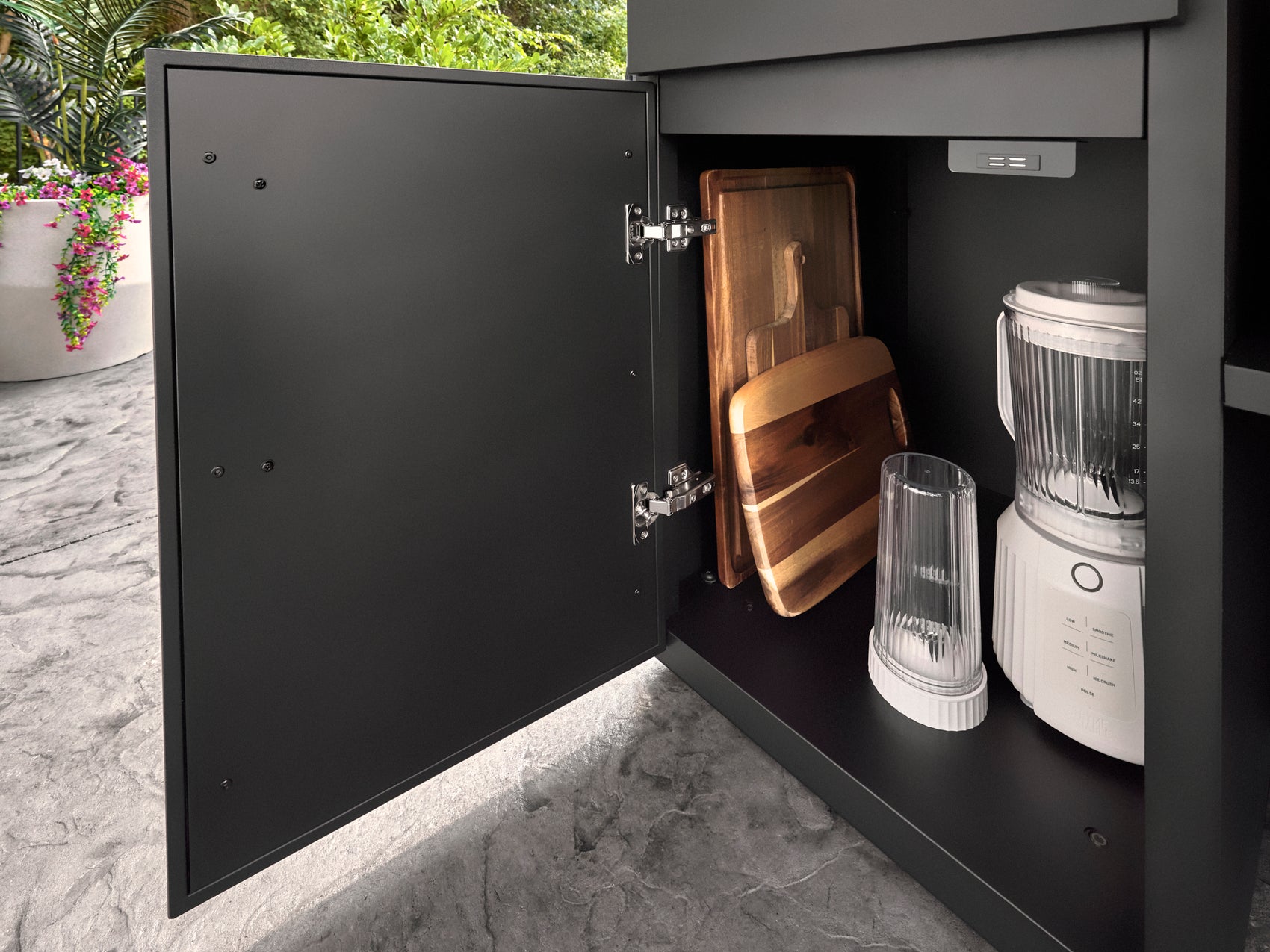 Lancaster powder-coated outdoor kitchen cabinet storing wooden cutting boards beside a blender on stone countertop.
