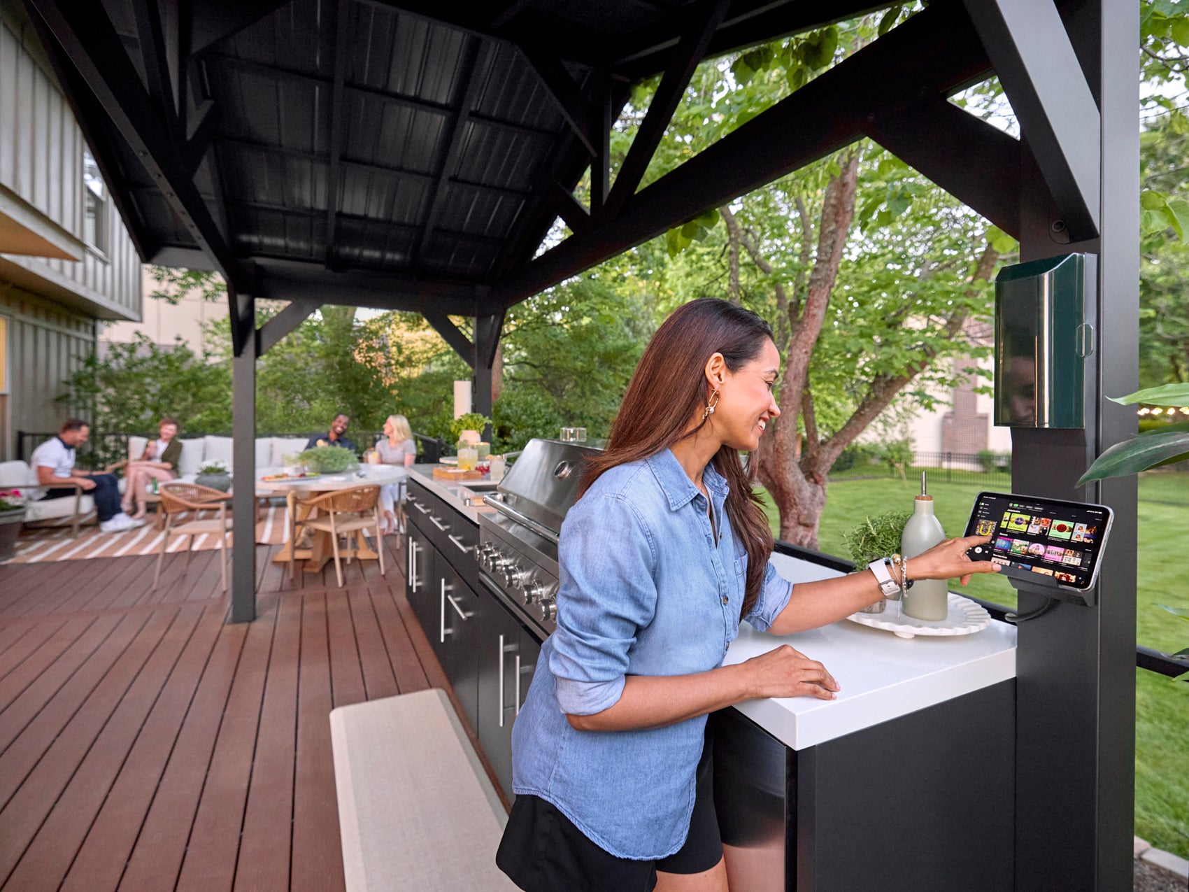 Woman using Lancaster outdoor kitchen with black steel roof and built-in grill during backyard gathering.