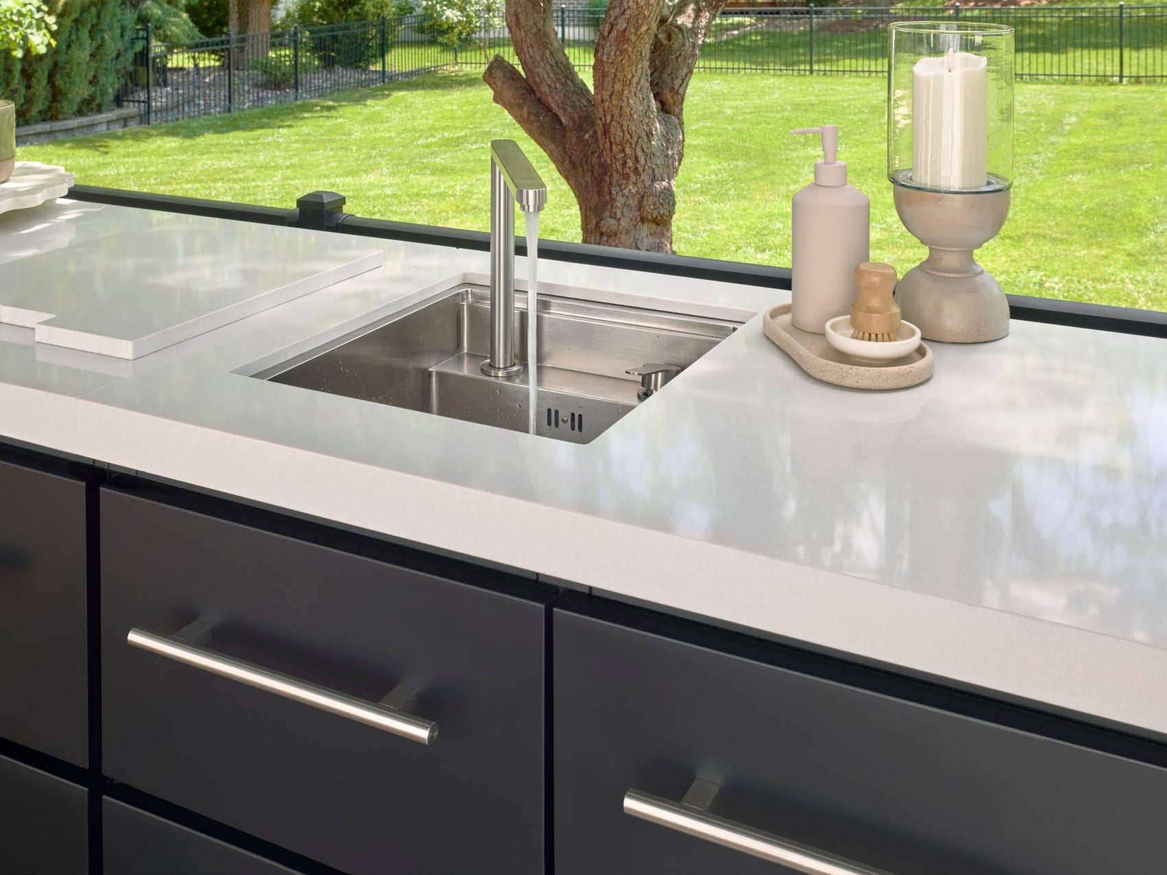 Modern kitchen with undermount sink and sleek faucet overlooking backyard pergola and manicured lawn through large window.