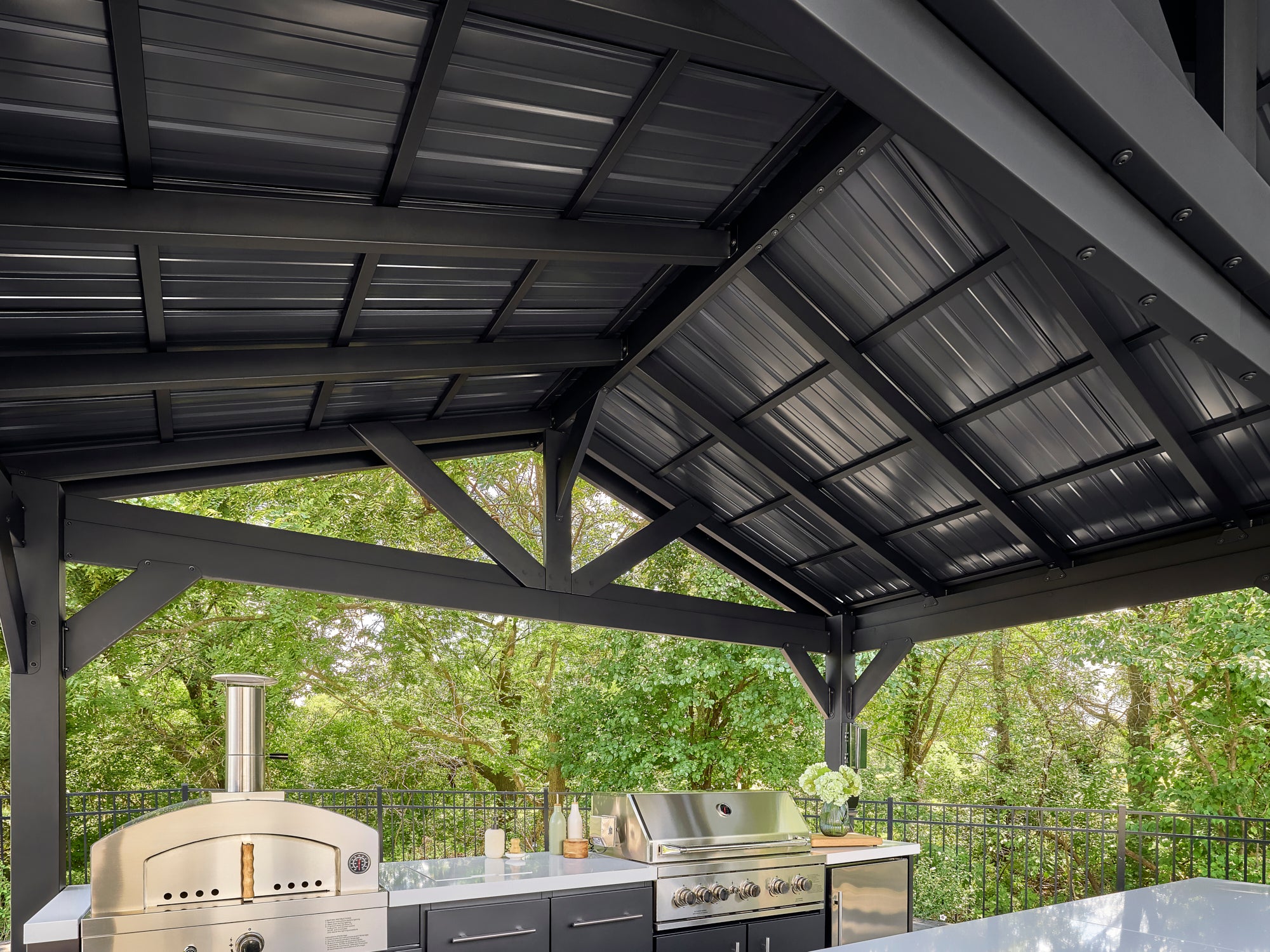 Powder coated galvanized steel roof of Lancaster outdoor kitchen with pizza oven and grill amid green trees.